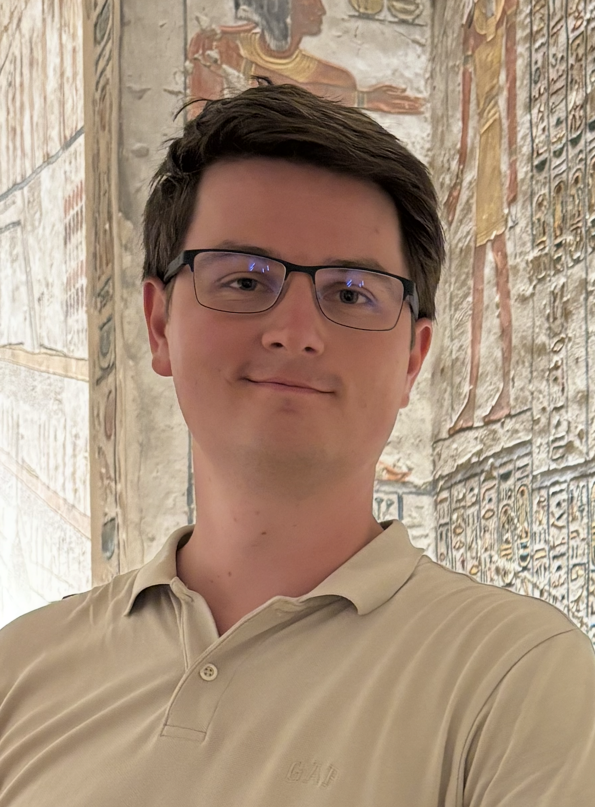 Muž v béžovém tričku stojí v chodbě s vytesanými egyptskými hieroglyfy na stěnách.